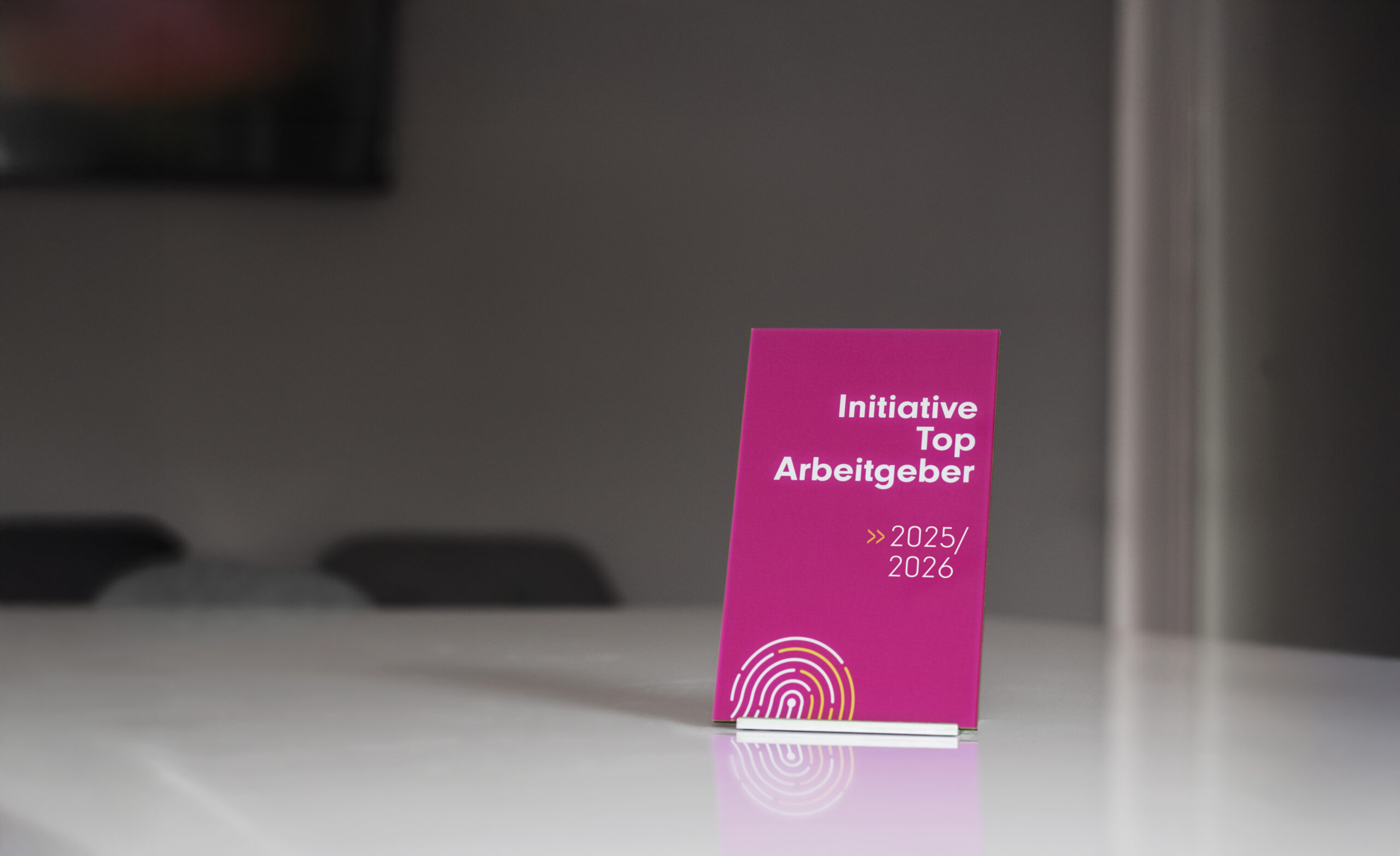 Auszeichnung „Initiative Top Arbeitgeber 2025/2026“ auf einem Tisch, präsentiert auf einer pinken Tafel vor unscharfem Hintergrund.