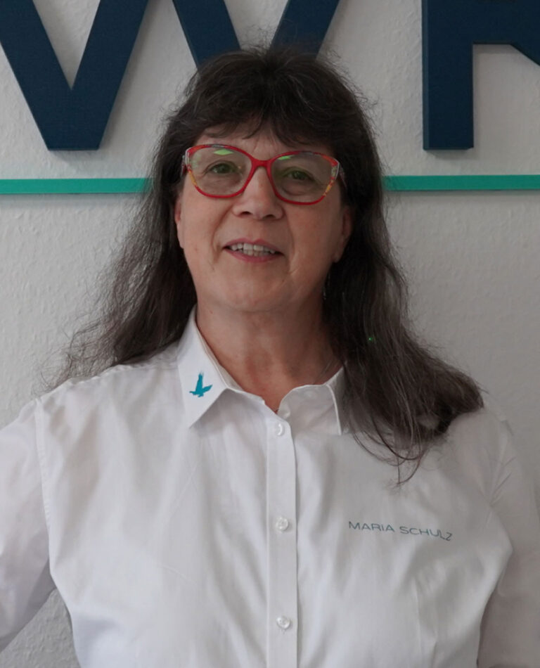 Porträt einer Frau mit roten Brillenrändern und weißem IPWK-Hemd, vor einer Wand mit IPWK-Logo.